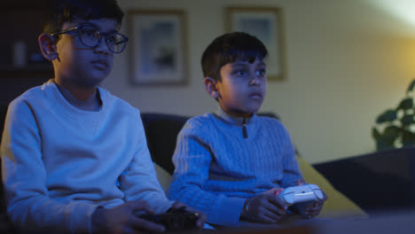 Two-Young-Boys-Sitting-On-Sofa-At-Home-Playing-With-Computer-Games-Console-On-TV-Holding-Controllers-Late-At-Night-5